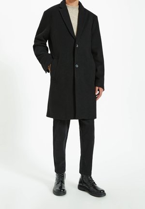 Cappotto di lana nera con rever a punta, due bottoni, tasche laterali, indossato su un maglione di cotone leggero, abbinato a pantaloni neri e stivaletti con lacci.