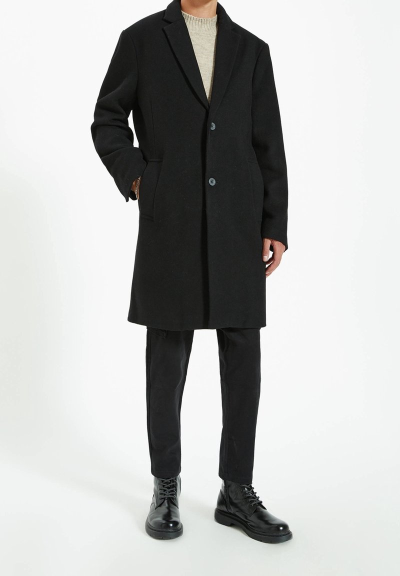 Cappotto di lana nera con rever a punta, due bottoni, tasche laterali, indossato su un maglione di cotone leggero, abbinato a pantaloni neri e stivaletti con lacci.
