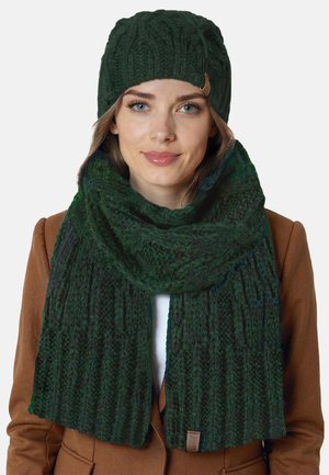 Mörkgrön stickad mössa med ribbad textur och liten detalj. Tillhörande mörkgrön scarf, även den ribbad, hänger runt halsen.