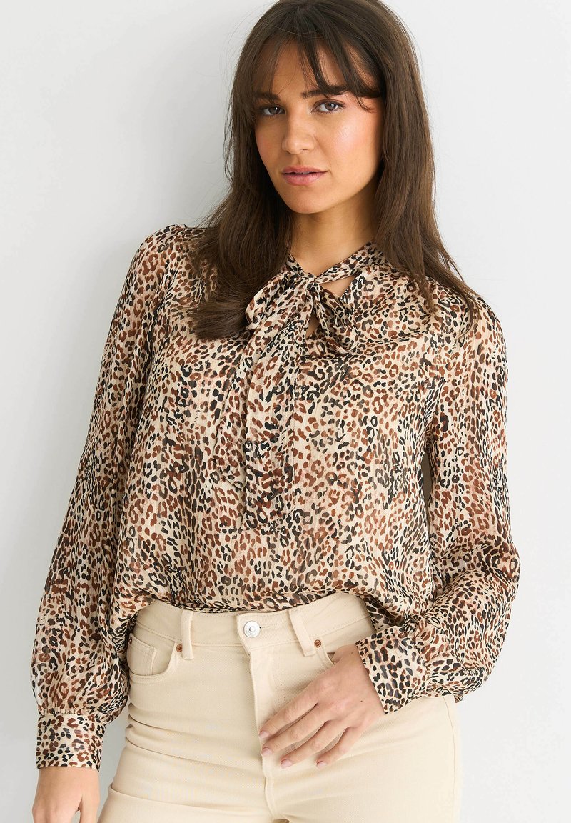 Leopard print blouse in lightweight fabric, featuring a tie at the neck and long sleeves with gathered cuffs. Paired with beige high-waisted pants.