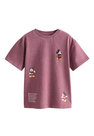 Junges Mädchen mit lockigem, zurückgebundenem Haar trägt ein lockeres lila T-Shirt und hellbeige Hose, steht mit dem Rücken zum Betrachter.