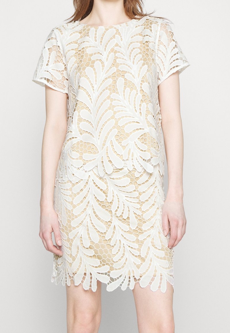 Femme portant une robe courte en dentelle blanche avec des motifs de feuilles et de nid d’abeille sur une sous-couche beige.