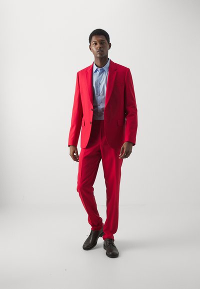Rød skræddersyet jakkesæt med én knaplukning, parret med en lyseblå skjorte. Mørke sko fuldender det formelle look mod en neutral baggrund.