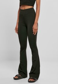 Leggings noires évasées en tissu extensible avec une taille haute. Texture lisse et design ajusté, accompagnés de sandales plates.