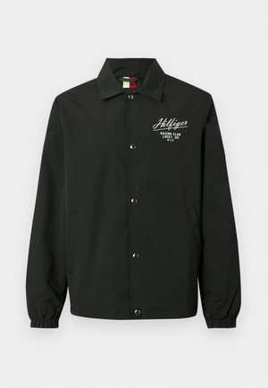 Mustapuku, joka on valmistettu kevyestä kankaasta, jossa on nappikiinnitys edessä, hihansuissa käänteet, sivutaskut ja valkoinen brodeerattu teksti rinnassa.