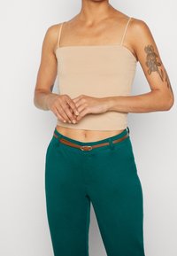 Béžová upnutá tílko s tenkými ramínky v kombinaci s tyrkysovými kalhotami s vysokým pasem. Hnědý opasek s kovovou sponou, hladké textury látky.