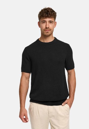 Mand med kort krøllet hår iført en sort kortærmet sweater og lyse beige bukser, stående foran en ensfarvet lys grå baggrund.