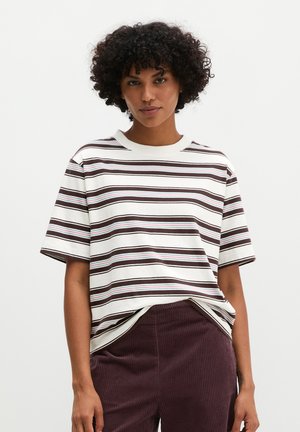 Vrouw met krullend haar die een wit shirt met korte mouwen draagt met bruine en blauwe strepen en een donkerbruine broek met hoge taille.