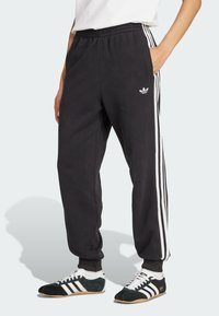 Crne fleece trenirke s bijelim bočnim prugama, elastičnim pojasom, bočnim džepovima i izvezenim logom na lijevom kuku.