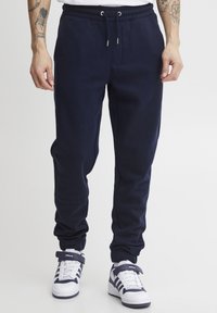 Navy blue sweatpants in soft fabric with a drawstring waistband, side pockets, and ribbed cuffs; paired with white sneakers.