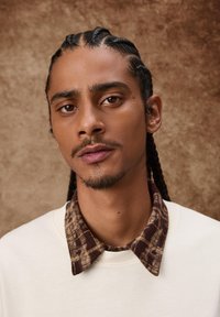 Lähikuva henkilöstä, jolla on päällään kermanvärinen collegepaita, jossa on ruskea ruudullinen kaulus. Henkilöllä on letitetty hiukset ja vaalea parta, taustana on teksturoitu pinta.