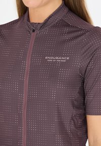 Maillot de cyclisme à manches courtes en violet foncé avec un petit motif à pois roses, fermeture éclair, et logo "ENDURANCE" sur le devant. Texture de tissu lisse.