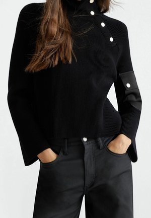 Femme portant un pull côtelé noir avec des boutons blancs au col et à la manche, les mains dans les poches d’un pantalon noir taille haute.