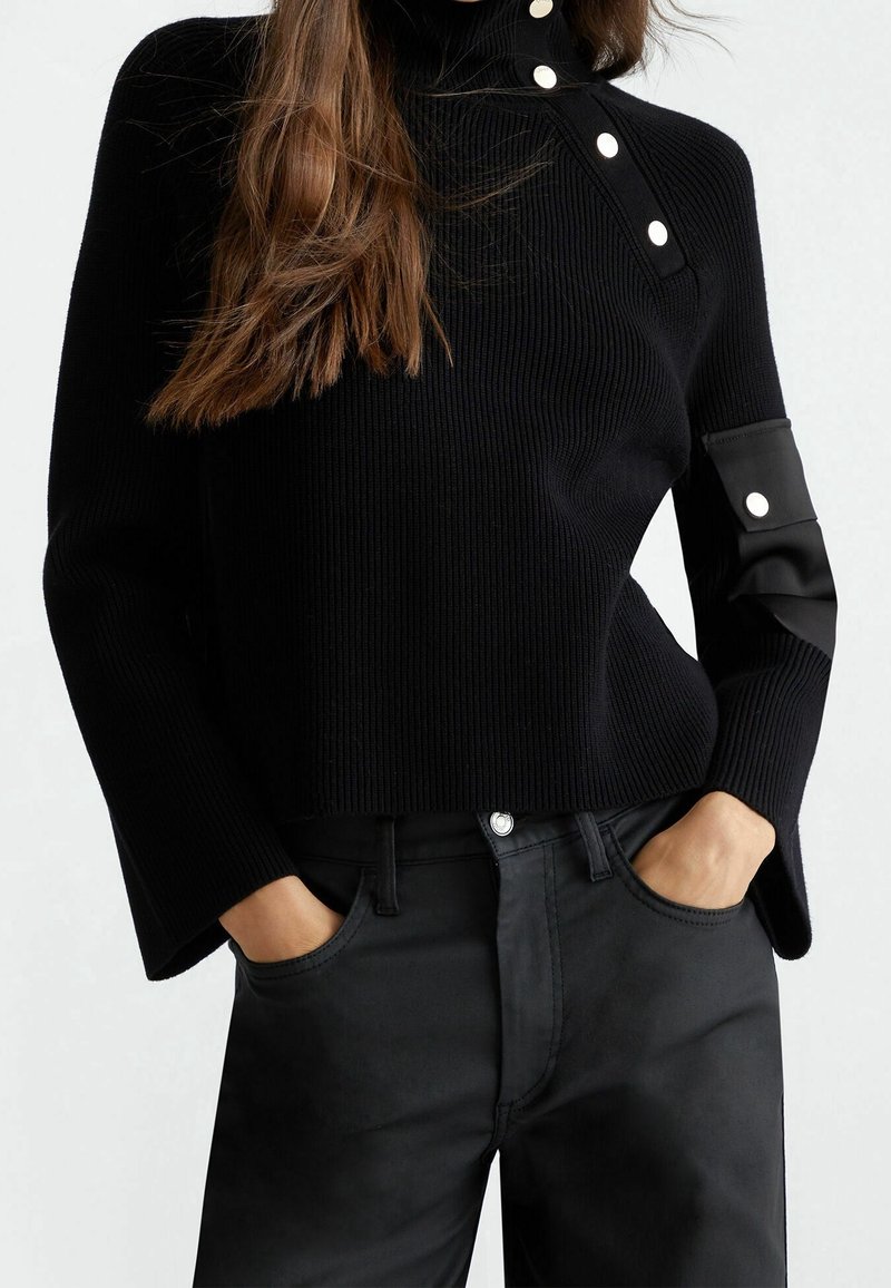 Femme portant un pull côtelé noir avec des boutons blancs au col et à la manche, les mains dans les poches d’un pantalon noir taille haute.