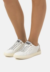 Zapatillas blancas con un upper de cuero texturizado, cordones plateados y detalles en tonos marrón en el talón, con una suela de goma de perfil bajo.