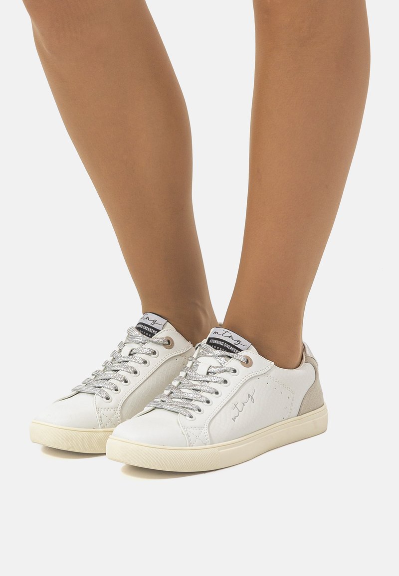 Zapatillas blancas con un upper de cuero texturizado, cordones plateados y detalles en tonos marrón en el talón, con una suela de goma de perfil bajo.