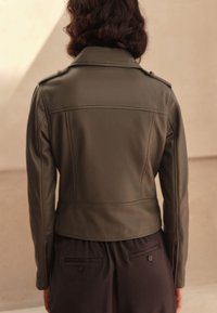 Giacca in ecopelle verde oliva con design aderente, spalline con bottoni e lunghezza corta. Texture liscia con dettagli a cucitura.