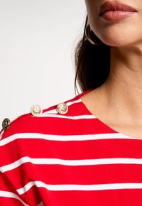 Rot-weißer gestreifter T-Shirt mit weitem Ausschnitt und vier dekorativen goldenen Knöpfen an der Schulter. Weicher Stoff.
