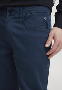 Mornarsko modre raztegljive hlače z gladko teksturo, ki imajo gumbasto zapiranje in stranske žepe. Majhen logotip blizu žepa.