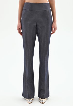 Persona che indossa pantaloni a vita alta, grigio scuro a righe sottili, abbinati a scarpe metallizzate con punta appuntita, in piedi su sfondo bianco.