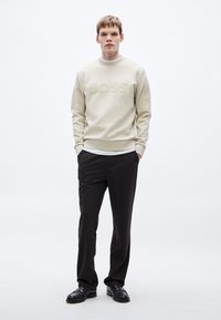 Μπεζ φούτερ με στρογγυλή λαιμόκοψη και ανάγλυφο λογότυπο "BOSS", συνδυασμένο με σκούρο παντελόνι και μαύρα παπούτσια, πάνω σε απλό λευκό φόντο.