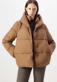Giacca a vento marrone con colletto alto e bottoni a pressione. Realizzata in tessuto trapuntato, presenta una silhouette ampia e una vestibilità morbida.