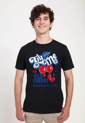 Usmívající se mladý muž s kudrnatými vlasy, oblečený v černém tričku s červenými třešněmi a textem propagujícím Wild Cherry Pepsi.