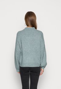 Donna con lunghi capelli castani che indossa un maglione a coste blu-grigio e pantaloni neri, di spalle, su uno sfondo chiaro e semplice.