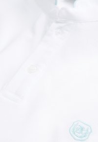Gros plan d'une chemise blanche à col avec un bouton et une petite rose brodée bleu clair près du bas à droite.