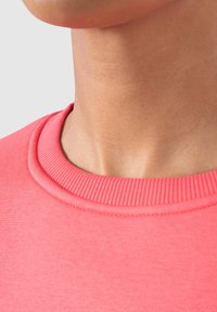 Korallrosa sweatshirt med ribbad rund halsringning. Slät yta, figurnära design som visar nacke och axelområde utan synliga sömmar.