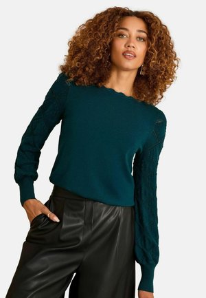 Türkisfarbener Strickpullover mit langen, strukturierten Ärmeln mit Rautenmuster und wellenförmigem Ausschnitt, kombiniert mit schwarzen Lederhosen.