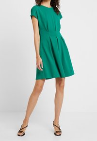 Grön kortärmad klänning med figursydd midja och utsvängd kjol; tillverkad av mjukt tyg med strukturerade veck och rund halsringning.
