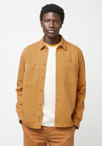 Braune Knopfjacke aus Baumwolle mit lockerer Passform, die zwei Brusttaschen und eine glatte Textur aufweist. Getragen über einem weißen Hemd.