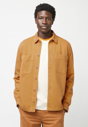 Bruine button-up jas van katoen met een ontspannen pasvorm, voorzien van twee borstzakken en een gladde structuur. Draagt over een wit shirt.