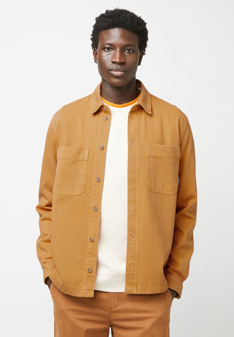 Braune Knopfjacke aus Baumwolle mit lockerer Passform, die zwei Brusttaschen und eine glatte Textur aufweist. Getragen über einem weißen Hemd.