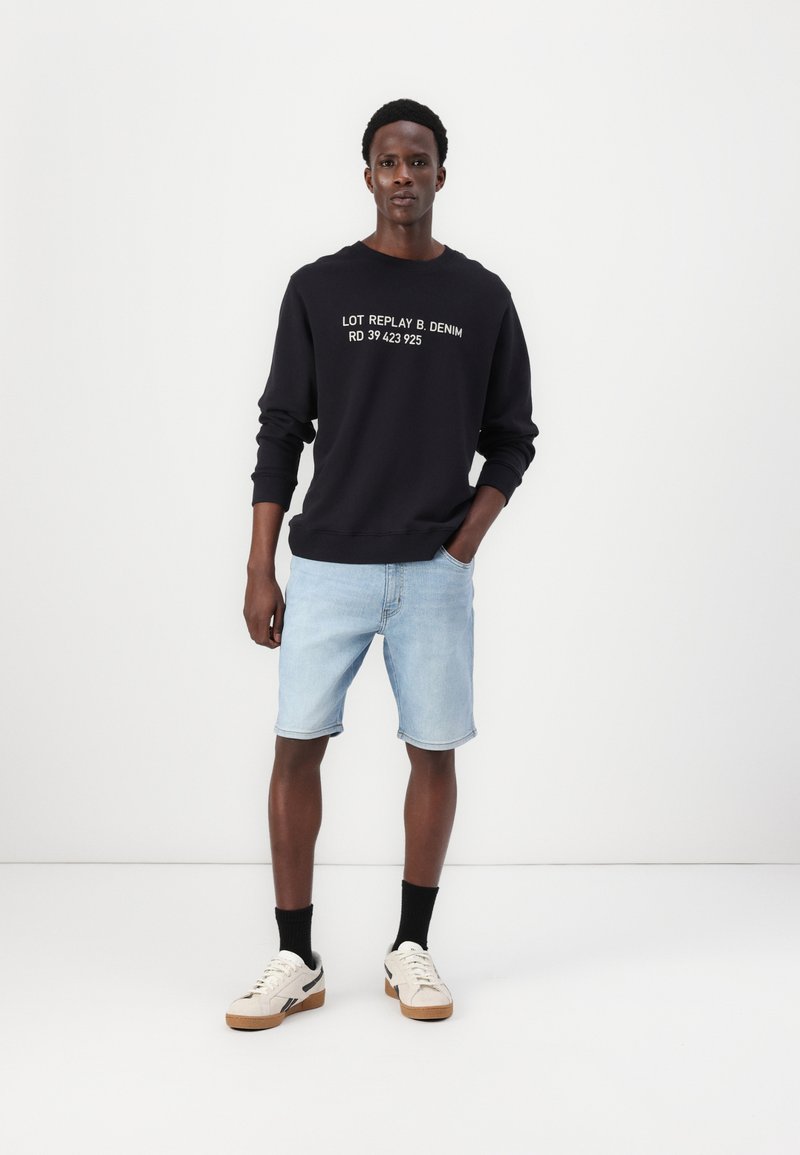 Sweat-shirt noir avec texte blanc, shorts en denim bleu clair et baskets beiges avec semelles noires. Fond uni, sans détails supplémentaires.