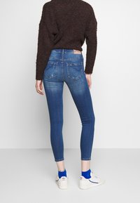 Vaqueros ajustados de color azul con cintura alta, tejido de mezclilla, desvanecimientos sutiles, bolsillos traseros y una etiqueta de marca marrón en la cinturilla. Zapatillas blancas con calcetines azules.