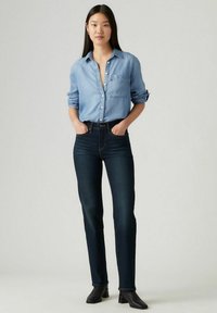 Camisa de mezclilla azul claro con un bolsillo en el pecho, cierre de botones en la parte delantera y mangas enrolladas. Jeans de tiro alto azul oscuro y botines negros. 