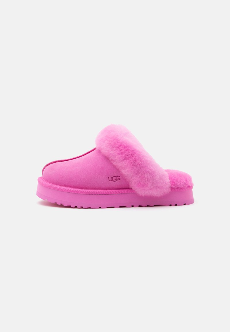 Φωτεινό ροζ παντόφλα τύπου slip-on με αφράτη γούνα στο εσωτερικό και σόλα, με το λογότυπο UGG στο πλάι, εμφανίζεται σε λευκό φόντο.