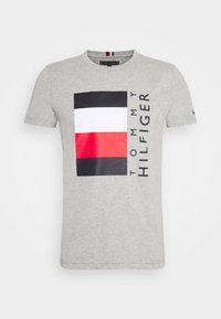 Camiseta de algodón gris con un gran gráfico de franjas horizontales en negro, blanco y rojo. Texto "TOMMY HILFIGER" impreso verticalmente.