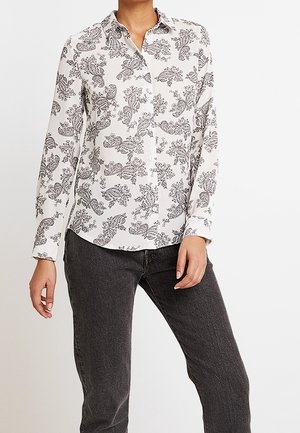 Nainen pukeutunut valkoiseen pitkähihaiseen nappipaitaan, jossa on musta paisley-kuvio, ja tummanharmaisiin farkkuihin, seisoo yksinkertaisen vaalean taustan edessä.