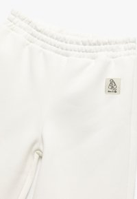 Pantalones blancos de cintura elástica con una pequeña etiqueta que muestra unos zapatos de ballet cruzados y la frase "la danza es vida" en la parte superior de la pierna.