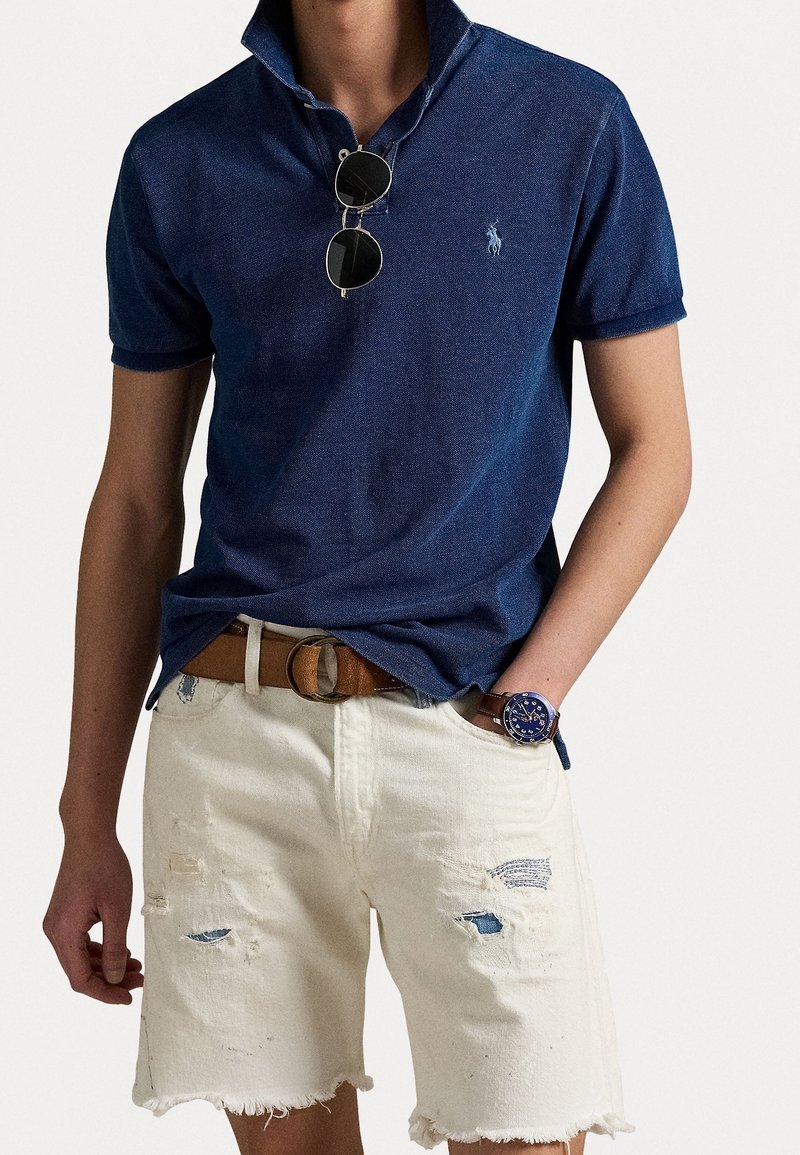 Personne portant un polo bleu foncé avec des lunettes de soleil suspendues au col et un short blanc usé avec une ceinture marron et une montre bleue.