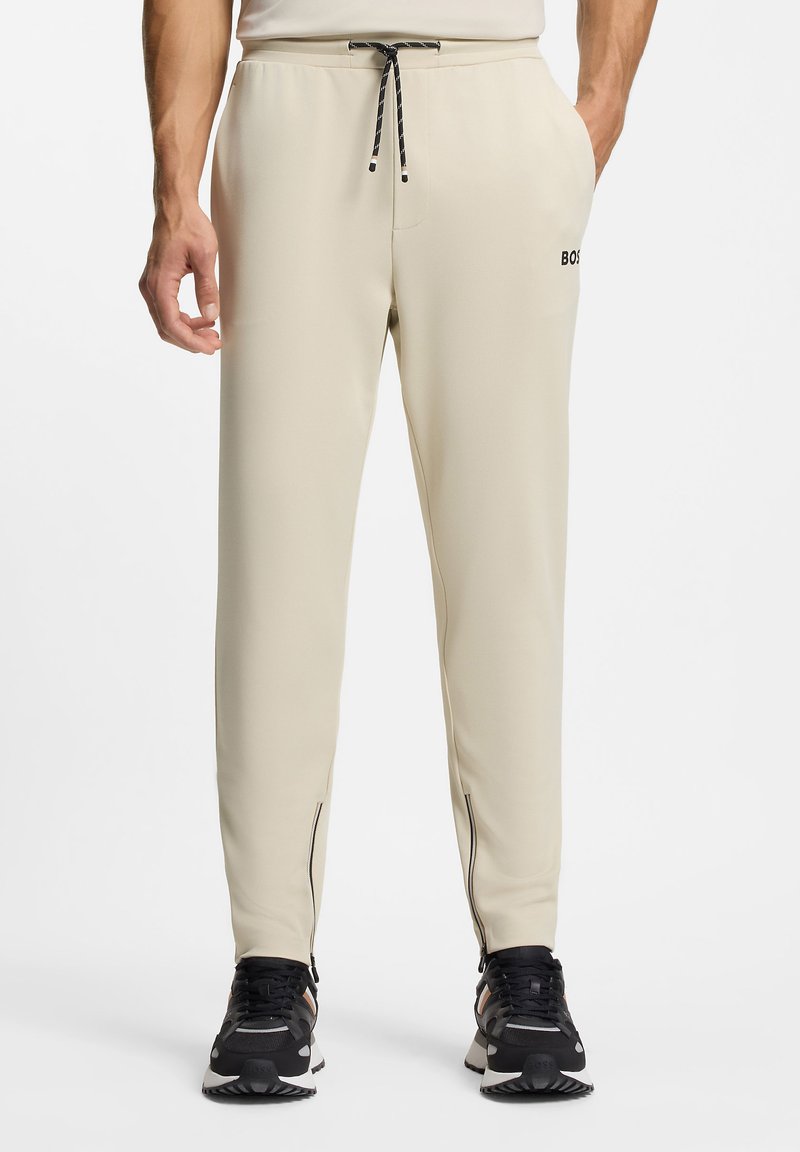 Beige athletic pants with a relaxed fit, featuring a drawstring waist, zippered hems, and logo accent. Smooth material and tapered design.