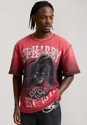 Hombre con cabello trenzado que lleva una camiseta roja de Ed Hardy con un diseño de pantera negra y flores, mirando hacia abajo con una mano en el bolsillo.