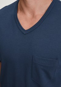 Close-up van een persoon die een marineblauwe V-hals T-shirt draagt met een klein zakje aan de linkerkant van de borst.