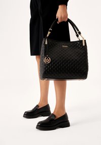 Borsa a tracolla in finta pelle nera con motivo impresso, hardware dorato e un charm decorativo. Presenta una forma strutturata e doppi manici.