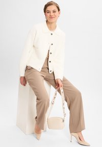 Junge Frau in beigen Hosen, weißer plüschiger Knopfjacke und beigen Heels, sitzt auf einem weißen Block und hält eine beigefarbene Umhängetasche.