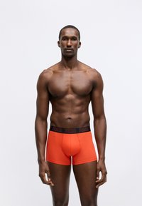 Red boxer briefs with a black logo waistband, made of smooth, stretchy fabric. The design is fitted, with a mid-rise cut.