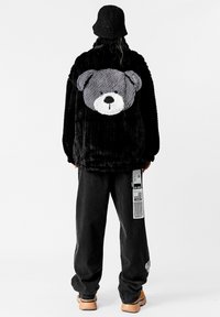 Chaqueta de piel sintética negra con un diseño de oso gris de peluche en la espalda, combinada con pantalones negros de corte holgado y un gorro de punto negro.